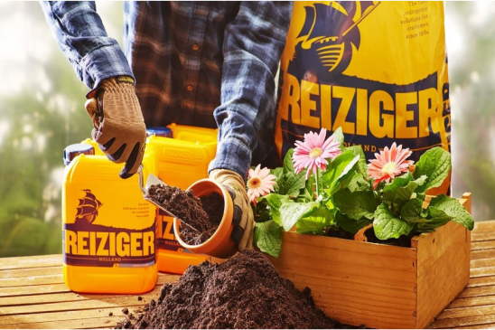 A partial view of a gardener in a plaid shirt and gloves, carefully pouring soil into a pot next to vibrant pink flowers in a wooden planter, with bright yellow 'REIZIGER' nutrient containers on a rustic wooden surface, set against a soft-focused natural backdrop.