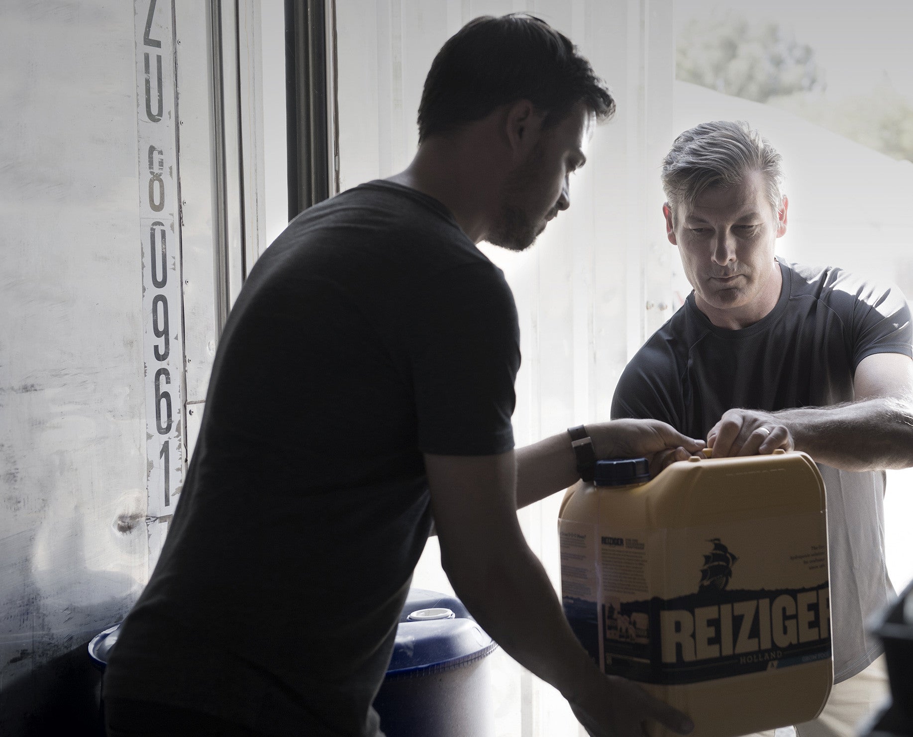 Two men working together indoors, one is passing a yellow industrial container to the other. There is a shipping container door in the background, suggesting an industrial or workshop setting